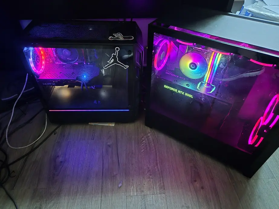 Two glass-panel gaming PCs side by side on a dark floor, lit in magenta and blue, with a GeForce RTX 3080 visible inside one